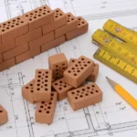 Mehrere Miniatur-Ziegelsteine, ein Bleistift und ein Zollstock sind auf der technischen Zeichnung eines Hauses angeordnet.