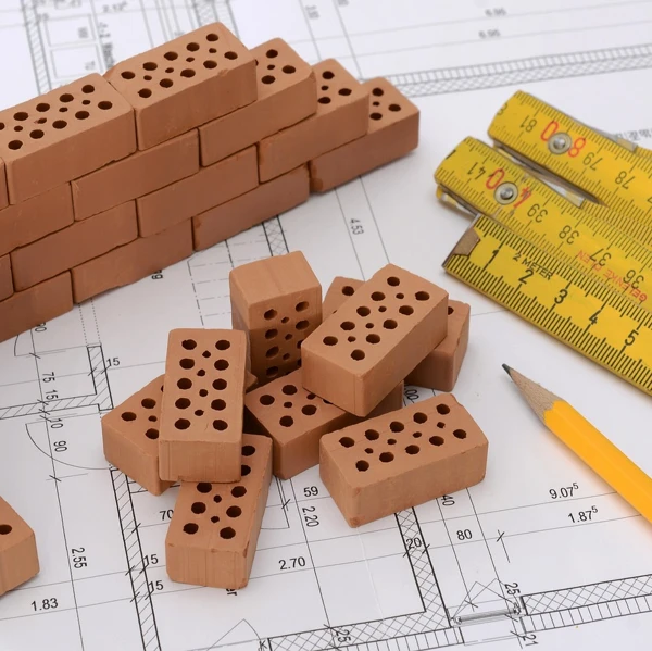 Mehrere Miniatur-Ziegelsteine, ein Bleistift und ein Zollstock sind auf der technischen Zeichnung eines Hauses angeordnet.